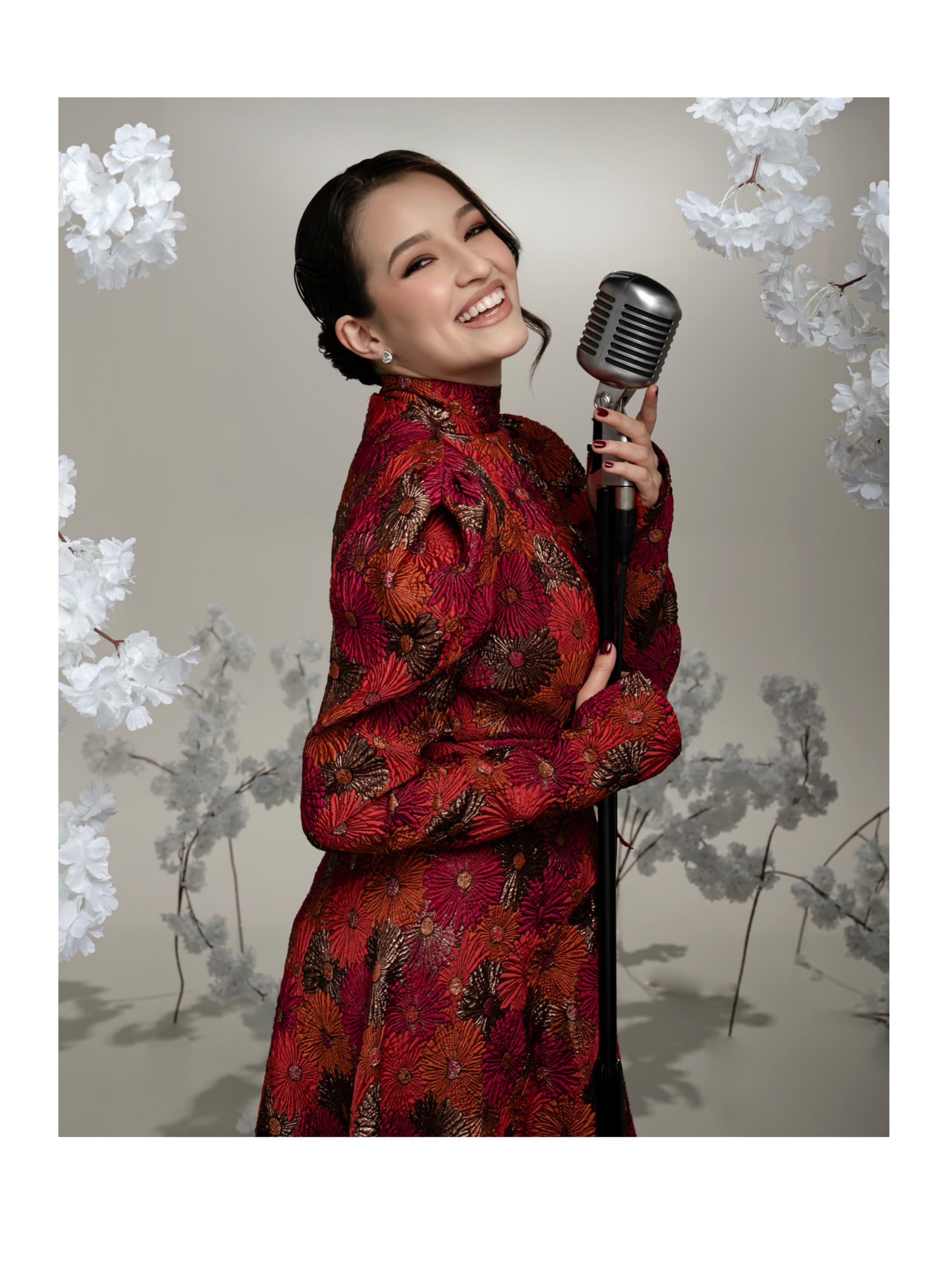 Jamilli Vaz com microfone vintage, em ensaio editorial entre flores brancas